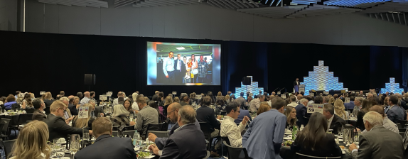 A room full of people at tables watching a video at the Site Selector Feedback dinner